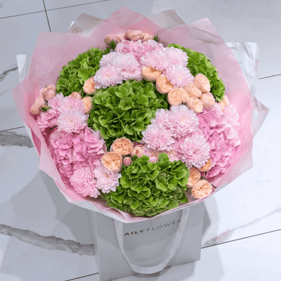 The “Wicked Blush” bouquet by Daily Flowers features pink and green hydrangeas, blush roses, pink chrysanthemums, and garden roses, elegantly wrapped in pale pink paper and presented in a white gift bag on a marble floor.
